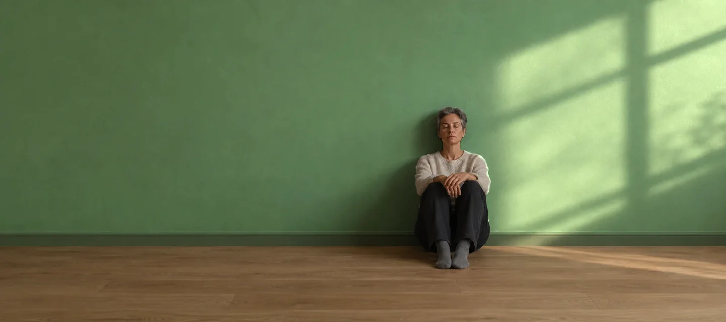 Burn-out : signes, causes et que faire Pause nécessaire après un burn-out : femme d'une cinquantaine d'années assise au sol contre un mur vert sauge, yeux fermés